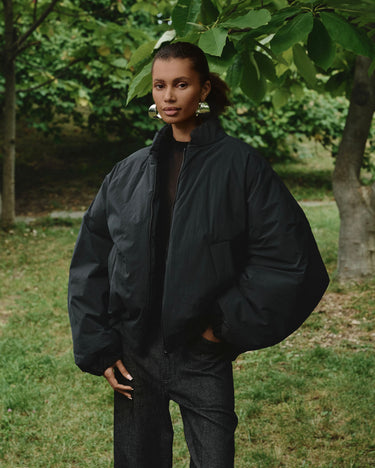 Padded bomber jacket, dark color with chest pocket detail, zip front, relaxed shoulders.
